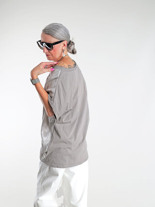 Frau trägt das OFF#DLY Tee Wake up taupe, ein lässig-elegantes T-Shirt mit Glanz-Front und Statement-Print, kombiniert mit Sonnenbrille. Vorderseite schimmert dezent, perfekt für modebewusste Outfits.