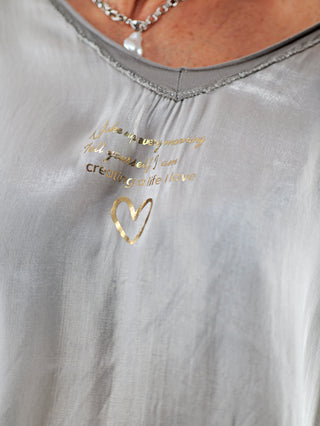 OFF#DLY Tee Wake up taupe: Nahaufnahme eines lässig-eleganten T-Shirts mit goldfarbenem Herz- und Statement-Print auf der schimmernden Vorderseite, kombiniert mit einem fließenden Stoffdesign.