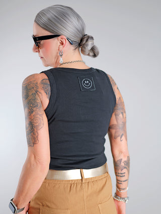 OFF#DLY Top Neve black: Frau mit Tattoos trägt schwarzes Baumwoll-Tanktop mit Smiley-Patch auf der Rückseite. Lässiger Schnitt, ideal für sommerliche Outfits, vielseitig kombinierbar. Einheitsgröße bis 40.