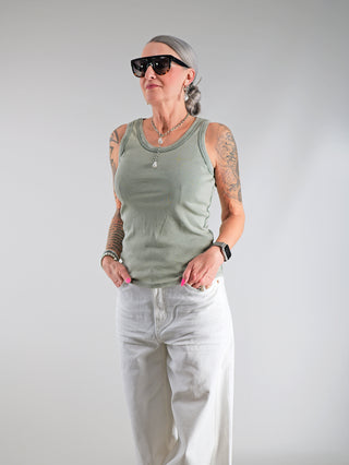Frau mit Tätowierungen trägt das OFF#DLY Top Neve khaki, ein lässiges Baumwoll-Top mit Smiley-Patch, Sonnenbrille und Armband, betont durch schmalen Schnitt und sportlichen Rundhalsausschnitt.