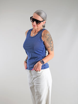 Frau mit Tattoos trägt das OFF#DLY Top Neve marine, ein lässiges Baumwoll-Tanktop mit Smiley-Patch, kombiniert mit weißen Hosen und großen Sonnenbrillen.