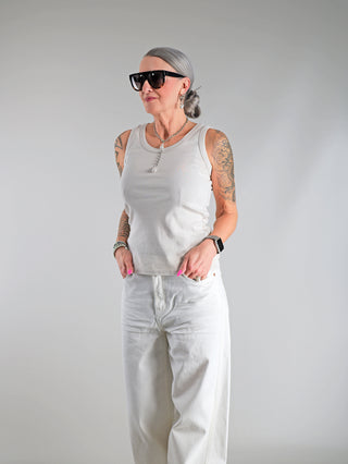 Frau trägt das OFF#DLY Top Neve sand, ein weißes Tanktop mit Smiley-Patch, kombiniert mit Sonnenbrille und weißen Hosen, betont durch Tattoos und pinke Nägel.