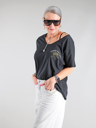 Frau trägt OFF#DLY Tee Angel blackgold, ein schwarzes T-Shirt mit goldfarbenem Engels-Print und positiver Botschaft, V-Ausschnitt, aus 100% Baumwolle, kombiniert mit Sonnenbrille und weißer Hose.