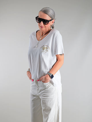OFF#DLY Tee Angel white/gold: Frau trägt weißes T-Shirt mit goldfarbenem Engels-Print und positiver Botschaft, V-Ausschnitt, lässige Passform, kombiniert mit Sonnenbrille, ideal für modischen Alltagslook.