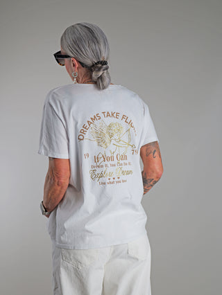 Frau trägt OFF#DLY Tee Angel white/gold mit goldfarbenem Engels-Print und Dreams take flight-Botschaft. Weißes T-Shirt mit V-Ausschnitt, lässiger Passform, aus 100% Baumwolle.