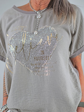 Person trägt das OFF#DLY Tee Believe taupe mit glitzerndem Paillettenherz und motivierendem Spruch. Das Shirt hat einen Rundhalsausschnitt und kurze Ärmel, passend bis Größe 40/42.