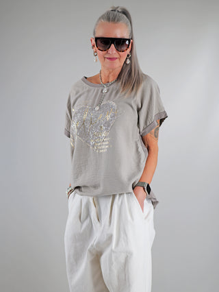 Frau trägt OFF#DLY Tee Believe taupe mit Paillettenherz und inspirierendem Spruch. Graues T-Shirt, lässiger Schnitt, Sonnenbrille und Ohrringe für einen modischen, femininen Look.