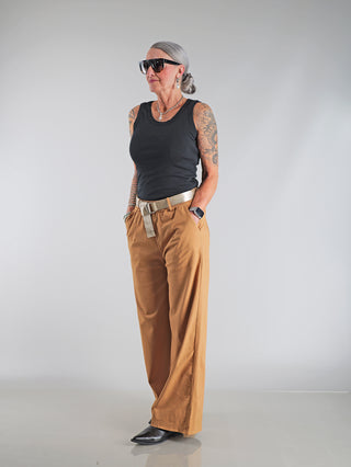 OFF#DLY Wide Leg Pant Rina camel: Frau mit Tattoos trägt weite Hose mit elastischem Bund und Glanzgürtel, betont durch stilvolle Coolness und schmeichelnde Silhouette.