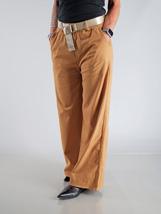 Person trägt OFF#DLY Wide Leg Pant Rina camel, weite Beine, markanter Glanzgürtel, elastischer Bund, seitliche und Gesäßtaschen. Elegante Silhouette mit bequemem Sitz. Perfekt für Alltag und Büro.