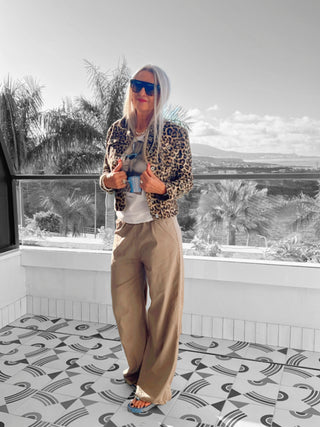 Frau trägt OFF#DLY Wide Leg Pant Rina taupe mit weitem Bein und elastischem Bund, kombiniert mit einem markanten Glanzgürtel, stehend vor Palmen im Hintergrund.