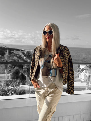 Frau auf Balkon mit OFF#DLY Tee Valencia weiß/blau, lässigem V-Ausschnitt und I don't care-Print, kombiniert mit einer Leopardenjacke und weißen Hosen, trägt weiße Sonnenbrille.