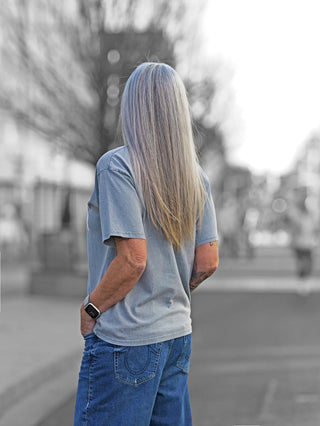Off#DLY Tee paris grey: Person mit langem Haar trägt graues Rundhals-T-Shirt mit PARIS-Print, kombiniert mit blauer Jeans, Uhr sichtbar am Handgelenk.