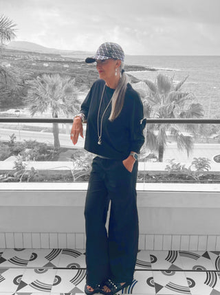 Frau steht vor Fenster und trägt OFF#DLY Wide Leg Pant Rina schwarz, kombiniert mit Sandalen und einem Hut.