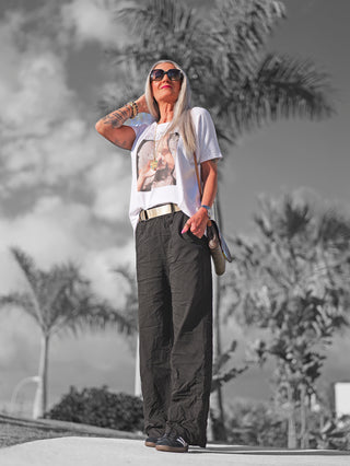Frau im Freien mit weiten OFF#DLY Wide Leg Pant Rina in Schwarz, kombiniert mit weißem Hemd und schwarzem Gürtel, umgeben von Palmen.