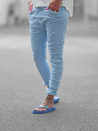 Eine Person trägt die OFF#DLY Pant Lynja lightblue, eine hellblaue Hose mit Rautenmuster und Crinkle-Look, kombiniert mit blauen und weißen Perlen-Sandalen.