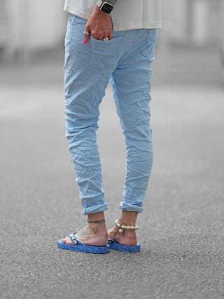 Beine einer Person in blauer OFF#DLY Pant Lynja lightblue, mit Crinkle-Look und Taschen, die lässig mit Händen in den Taschen steht und Sandalen trägt.