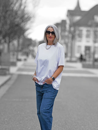 Nuomeno Oversized Tee Nube: Frau in weißem, locker geschnittenem Baumwoll-T-Shirt mit rückseitigem Print, kombiniert mit blauer Jeans, steht auf einer Straße und trägt Sonnenbrille.