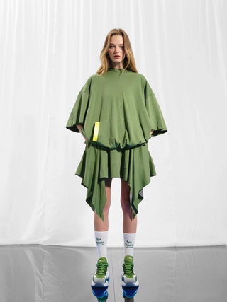 #VDR Sweater oliv: Frau trägt grünes, oversized Sweater mit Kapuze, asymmetrischem Saum und Pailletten-Backprint. Kombiniert mit grünen und weißen Sneakers.
