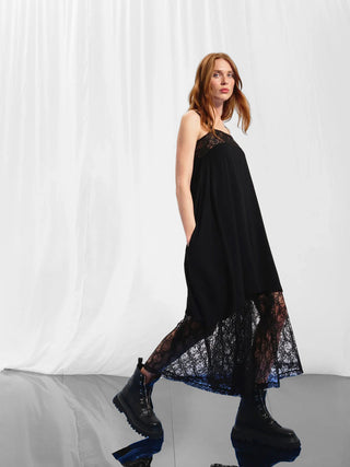 Frau trägt #VDR Jumpsuit nero mit Spitzeneinsatz, fließender Silhouette und weiten Beinen, kombiniert mit schwarzen Boots. Elegantes Design mit femininer Note für vielseitige Looks.