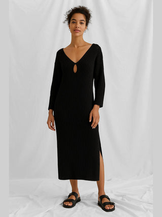 Frau trägt das Cotton Candy Nelly Dress black, ein Maxikleid mit seitlichen Schlitzen und Dekolleté-Cut-Outs, kombiniert mit schwarzen Sandalen, ideal für sommerliche Eleganz.
