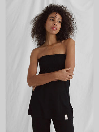 Frau in schwarzem Cotton Candy Nica Top mit gesmoktem Bund und Seitenschlitzen, schulterfrei und elegant, trägt goldene Kette, Modellgröße 1,76 m. Stoff: 55% Viskose, 45% Polyester.
