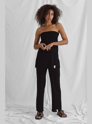 Eine Frau trägt das Cotton Candy Nica Top schwarz mit gesmoktem Bund und Seitenschlitzen, kombiniert mit einer schwarzen Hose.