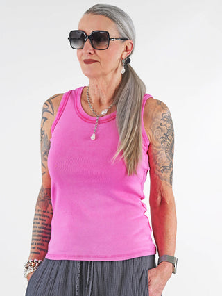 Frau mit Tattoos trägt das OFF#DLY Top Neve pink, ein ärmelloses Baumwoll-Top mit Smiley-Patch, kombiniert mit Sonnenbrille und Armbanduhr, ideal für einen lässigen Sommerlook.
