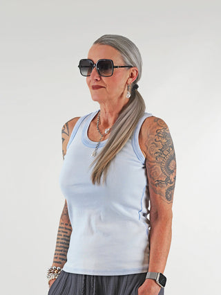 Frau mit Sonnenbrille trägt das OFF#DLY Top Neve lightblue, ein ärmelloses Baumwoll-Top mit Smiley-Patch, das lässig und vielseitig kombinierbar ist. Perfekt für Sommeroutfits.