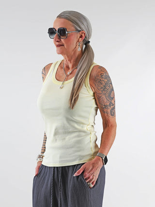 Frau mit tätowiertem Arm trägt das OFF#DLY Top Neve gelb, ein lässiges Baumwoll-Tanktop mit Smiley-Patch im stilvollen Design, kombiniert mit Sonnenbrille und Accessoires.