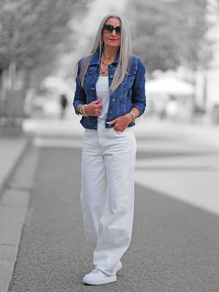 Ältere Frau trägt die OFF#DLY Denim Jacket dark blue, tailliert und stilvoll, über einem weißen Tanktop, kombiniert mit weißen Jeans und Sneakers auf einer gepflasterten Stadtstraße.