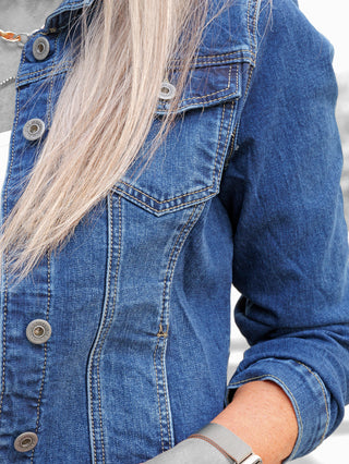 Nahaufnahme einer Person, die die OFF#DLY Denim Jacket dark blue trägt, mit silbernen Druckknöpfen, einem silbernen Kettenhalsband und einer Metall-Uhrenarmband, lange blonde Haare fallen über die dunkelblaue Jacke.
