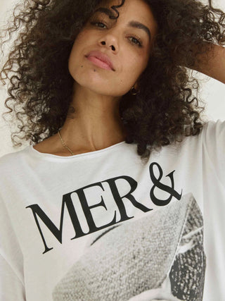 Eine Frau mit lockigem Haar trägt das Cotton Candy Tee Helin white mit MER & SOLEIL Aufdruck und 3/4 Ärmeln, betont durch einen nahen Blick auf das T-Shirt.
