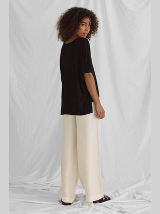 Frau trägt das Cotton Candy Tee Helay in Schwarz mit V-Ausschnitt und Saumversatz, kombiniert mit weißen Hosen. Das T-Shirt ist oversized und aus Viskose-Polyester-Mischung.