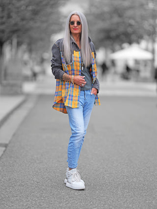 Frau auf Straße trägt OFF#DLY Blusenjacke Denim mit grauem Denim-Oberteil und gelb-blauem Karomuster unten, Ziernieten an Brusttaschen, lässiger Schnitt.