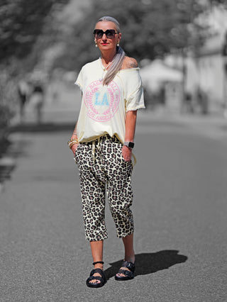 Frau trägt OFF#DLY Leo Short gelb, eine lässige Hose mit auffälligem Leo-Print und elastischem Bund, kombiniert mit schwarzen Sandalen und weißem Shirt mit pinkem Logo auf der Straße.