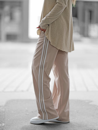 Person trägt OFF#DLY Pant Stripes beige/white mit lockerer Passform und weißen Streifen. Kombiniert mit weißen Slip-on-Sneakers und einem langen Pullover, betont die Hose einen sportlich-eleganten Look.