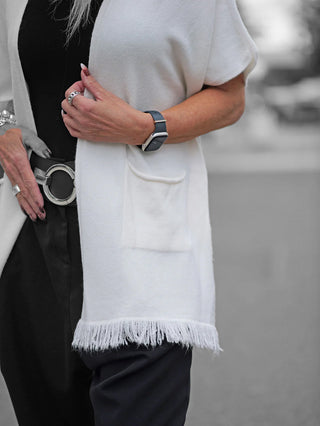 Nahaufnahme einer Frau mit der OFF#DLY Strickjacke Sabrina creme, offen getragen über einem schwarzen Outfit. Die Strickjacke hat Fransen am Bund und wird mit einem breiten schwarzen Gürtel akzentuiert.