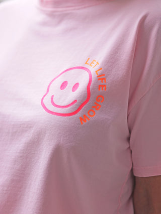 Person trägt das OFF#DLY Tee Smile rosa mit kleinem Smiley-Print vorne und inspirierendem Design hinten. Das T-Shirt wirkt weich und dehnbar, ideal für lässige Freizeit-Outfits.