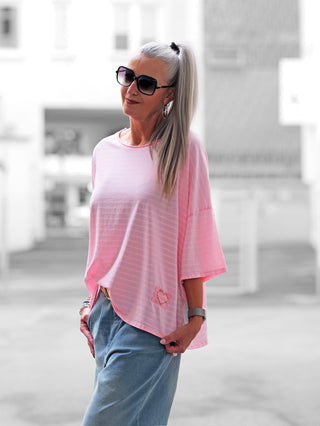 Eine Frau trägt das OFF#DLY Stripes Shirt rosa/pink mit Herz-Print am Saum. Das T-Shirt ist lässig, hat Streifen und eine bequeme Passform, ideal für Freizeitoutfits.