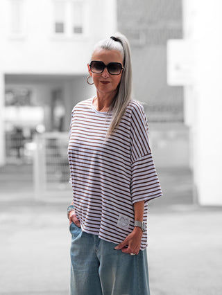 Frau trägt das OFF#DLY Stripes Shirt in Braun/Weiß, gestreifter Stil mit Herz-Print „l’amore è bello“ am Saum, kombiniert mit Jeans, passend für Freizeit und Sommer.