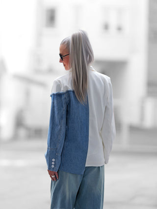Person mit silbernem Pferdeschwanz, Sonnenbrille und dem OFF#DLY Blazer Lazise white denim, der aus weißem Stoff und blauem Denim besteht, ergänzt durch passende weite Denim-Hosen.