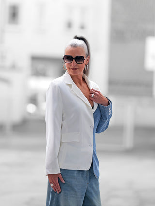 Frau mit silbergrauem Pferdeschwanz trägt den OFF#DLY Blazer Lazise white denim, asymmetrisch zweifarbig, kombiniert mit locker sitzenden Jeans, silbernen Accessoires und dunklen Sonnenbrillen, steht leicht lächelnd im urbanen Hintergrund.