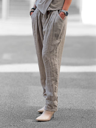 Person trägt die OFF#DLY Pant Volterra taupe, eine locker geschnittene Leinenhose mit Bundfalten und geflochtenem Gürtel, passend für warme Tage. Ideal kombinierbar mit verschiedenen Schuhen.