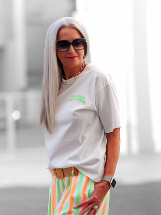 Nahaufnahme einer Frau mit Sonnenbrille trägt das OFF#DLY Tee LA 91 in offwhite, ein weißes T-Shirt mit sportlichem Neonprint, das urbane Westcoast-Vibes verkörpert.