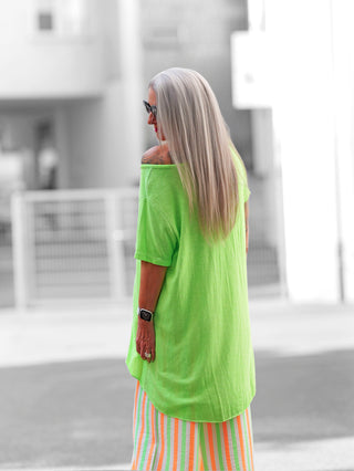 Eine Frau trägt das OFF#DLY Longshirt Laglio neongrün mit asymmetrischem Saum, lockerem Oversize-Fit und Off-Shoulder-Option, ideal für lässige Sommerlooks.