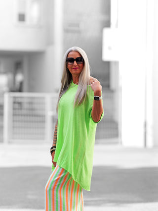 Frau trägt das OFF#DLY Longshirt Laglio neongrün mit lässigem, asymmetrischem Schnitt. Kombiniert mit einer orange-weiß gestreiften Rock für einen modernen Street-Fashion-Look.
