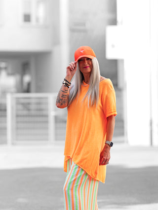 Das OFF#DLY Longshirt Laglio neonorange zeigt einen lässigen Oversize-Schnitt mit asymmetrischem Saum, getragen von einer tätowierten Person, die das modische Shirt als stilvollen Sommerlook präsentiert.