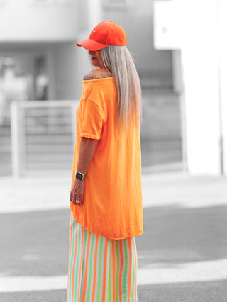 Person trägt OFF#DLY Longshirt Laglio neonorange mit lässigem Oversize-Fit und asymmetrischem Saum, ideal für sommerliche Anlässe, abgerundet durch einen modischen, über die Schulter tragbaren Schnitt.