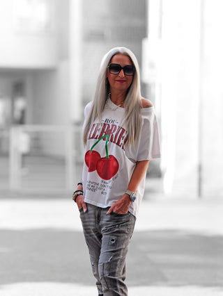 Frau trägt das OFF#DLY Tee Cherry white, ein weißes T-Shirt mit roten Kirschen-Print, lässigem Schnitt und weitem Rundhalsausschnitt, ideal für entspannte Sommertage.