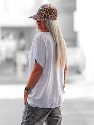 Person von hinten in OFF#DLY Tee Heart min min white, locker sitzendes weißes T-Shirt mit goldenem Herzprint, kombiniert mit grauer Jeans und Leoparden-Basecap; zeigt langen Haaren und Smartwatch.
